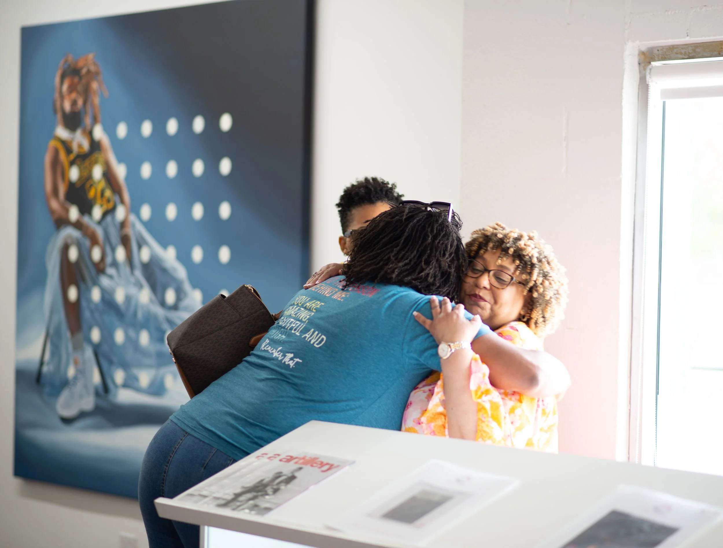 Two women hugging each other in an art gallery or museum, with a colorful artwork on the wall behind them and a window on the right.