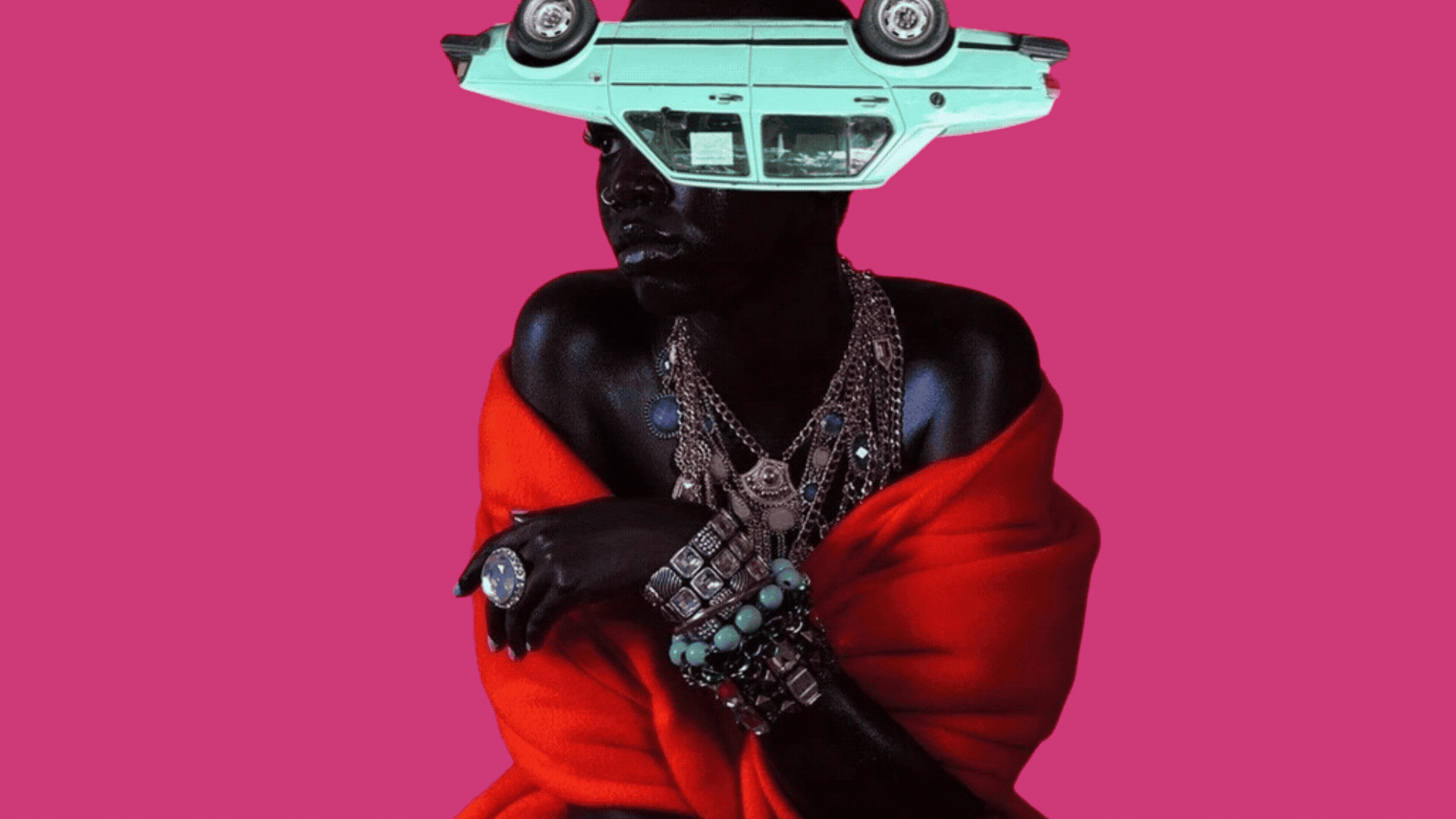 A person with dark skin and multiple jewelry pieces, including necklaces, rings, and bracelets, is wrapped in a red fabric against a pink background. Their face is partially obscured by a vintage turquoise toy car worn upside down on their head.
