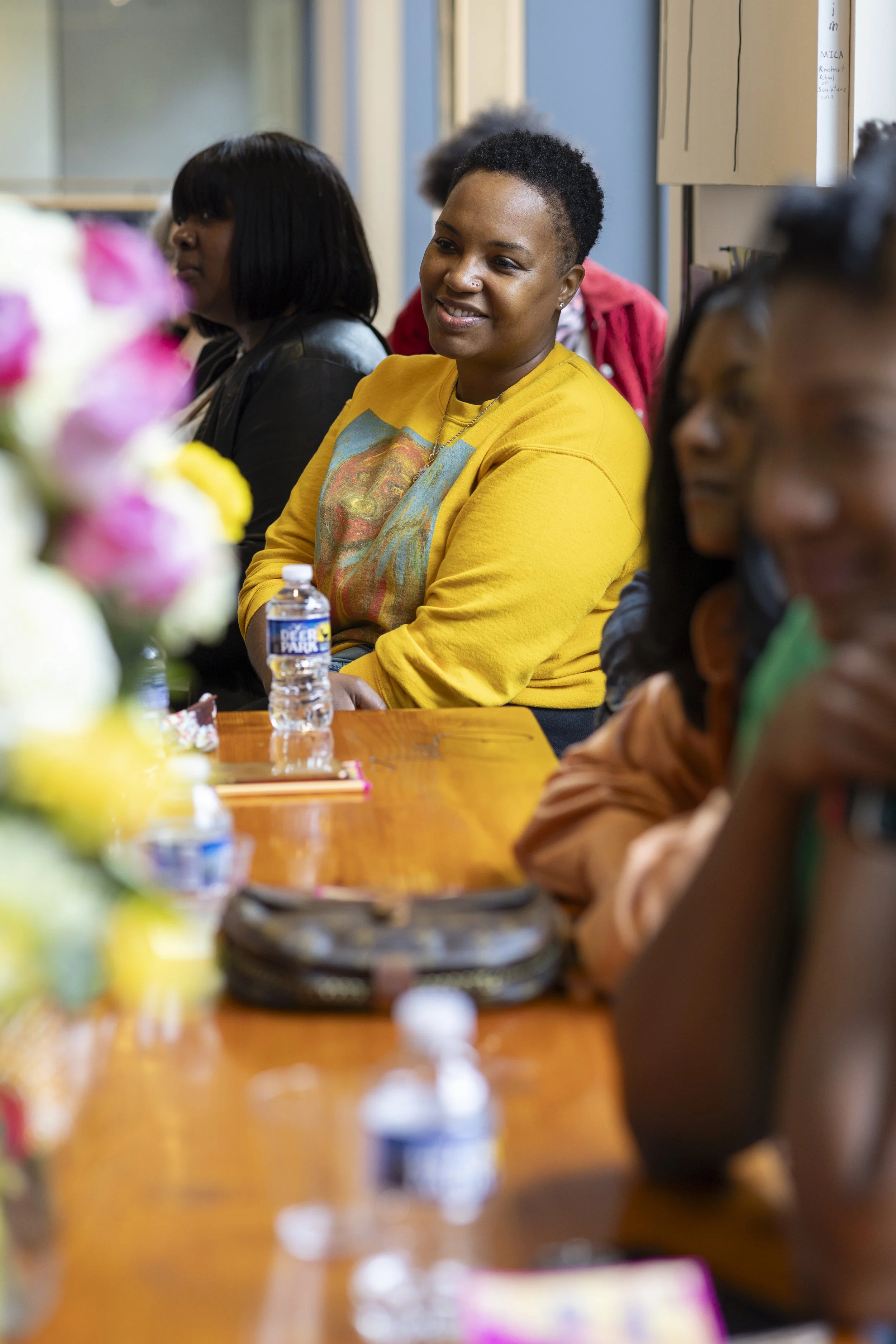 A woman wearing a yellow sweatshirt with a graphic design, sitting at a table with water bottles and flowers, smiling and listening attentively during a meeting or event inside a room.