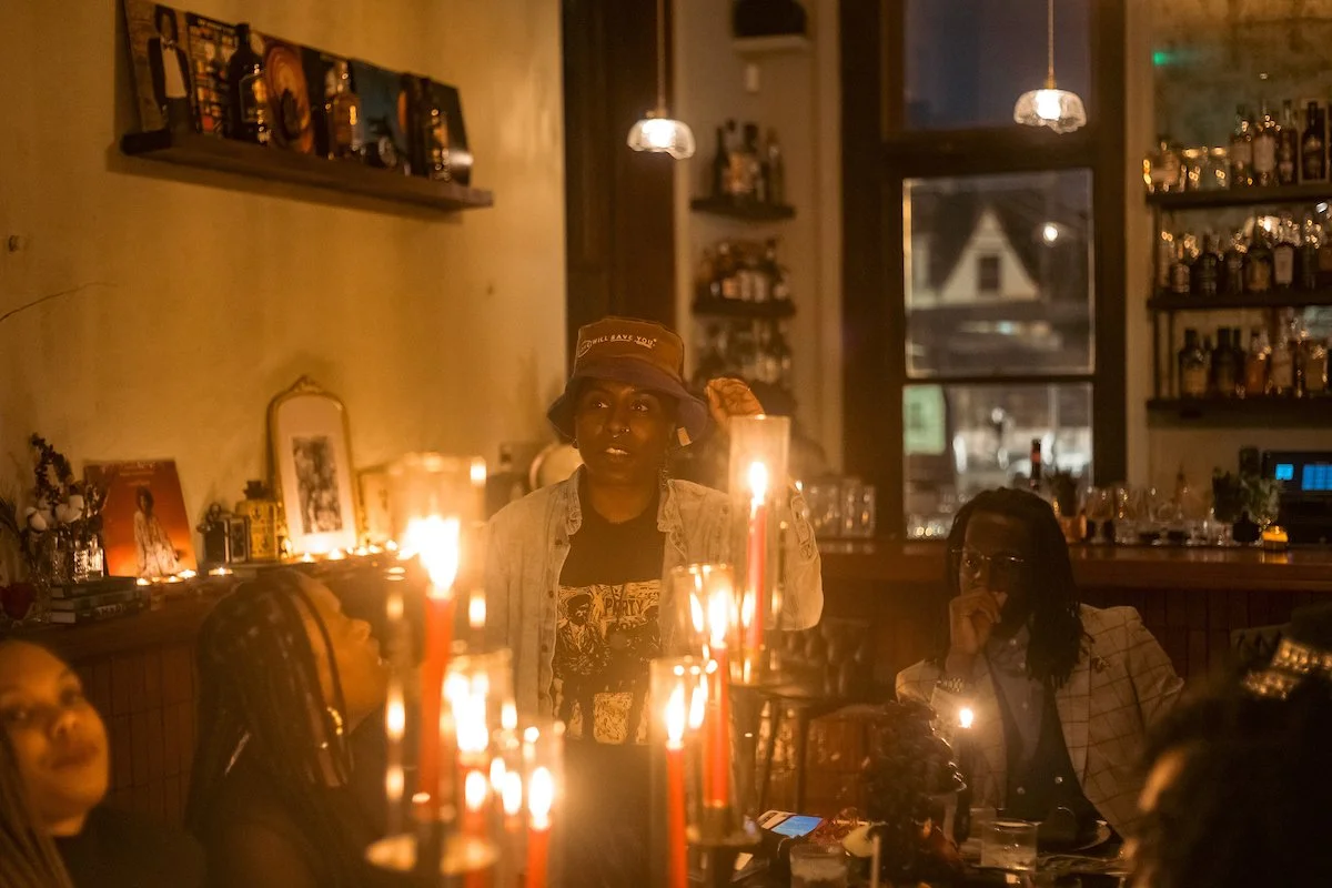 People gathered in a dimly lit restaurant or bar with candles on the table, shelves with bottles and pictures on the wall, and a window in the background. A woman in the center wears a bucket hat and denim jacket, looking attentively.