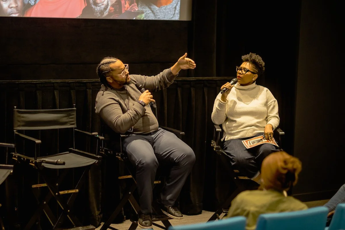 Two people speaking into microphones on stage, seated in black director's chairs, engaged in conversation during an event or panel discussion, with an audience member visible in the foreground.