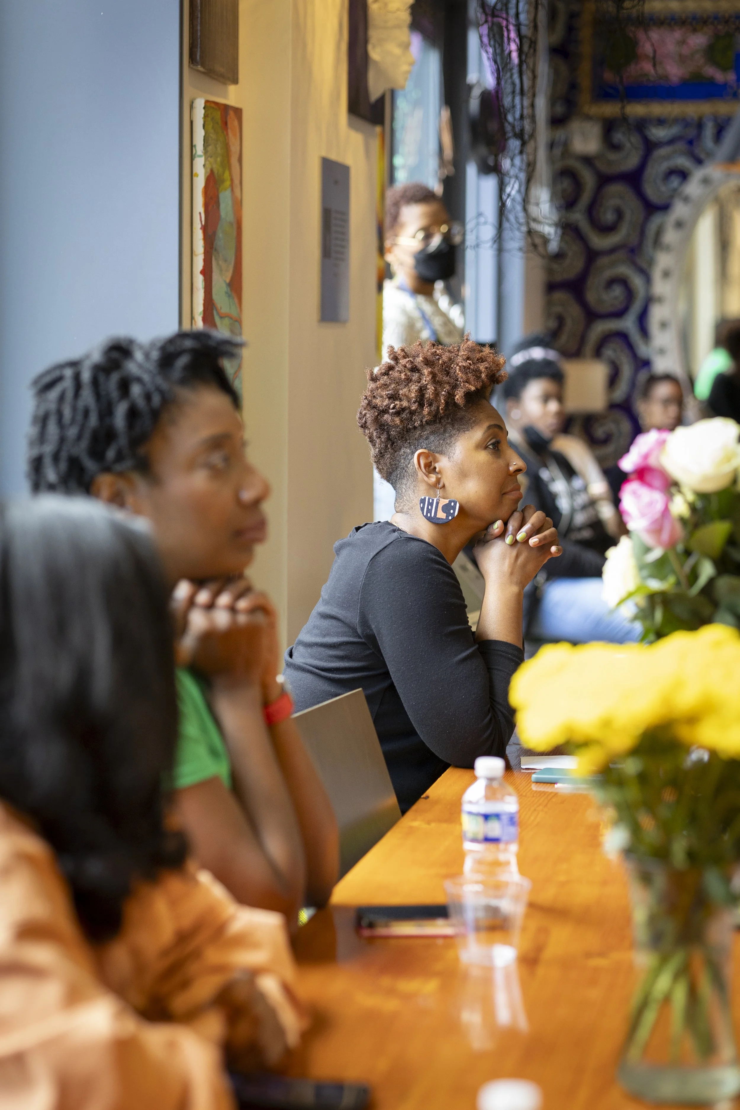 Group of diverse women listening attentively during a meeting or seminar in a well-lit room with artistic wall decorations and a table with flowers and water bottles.