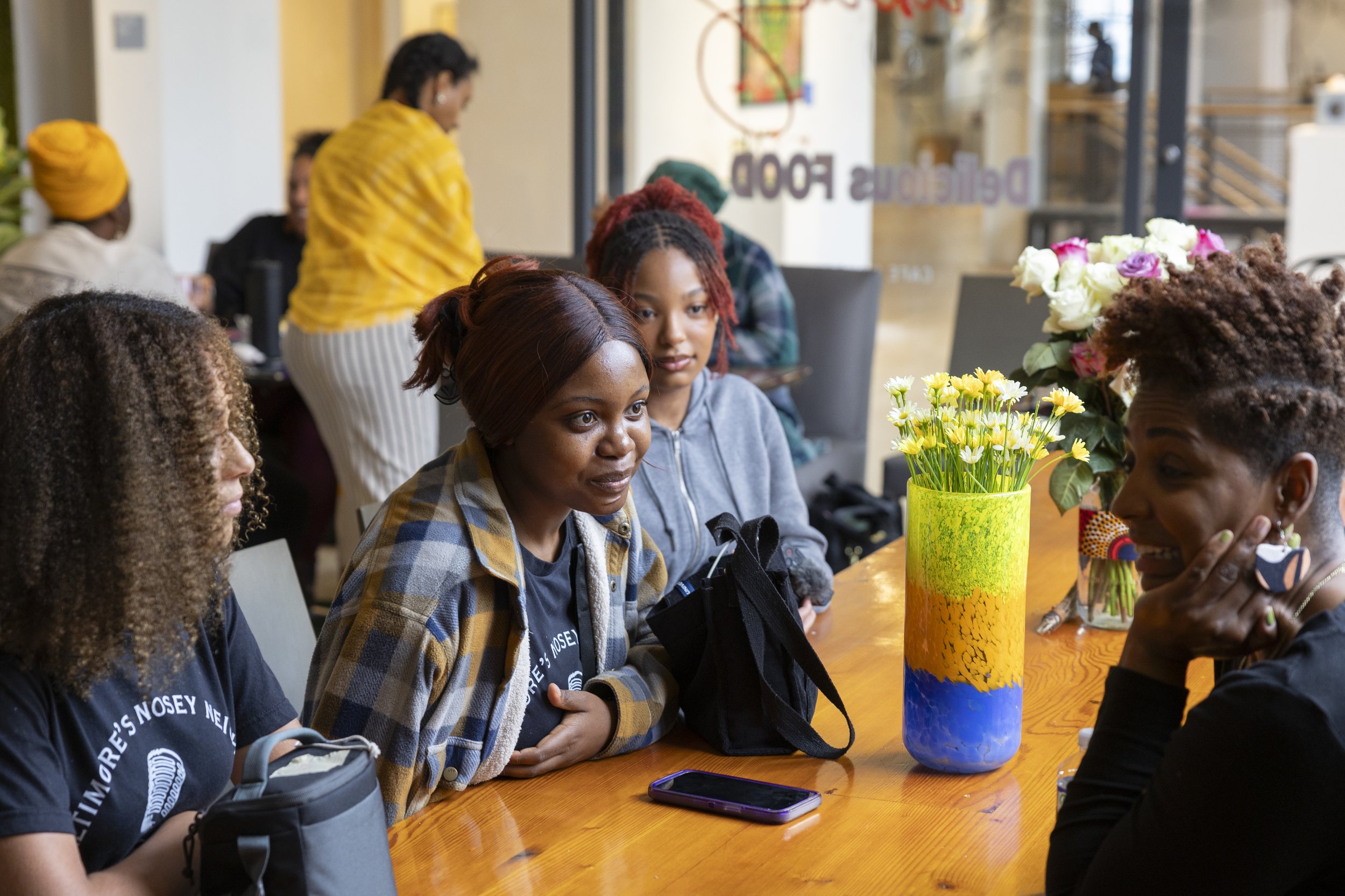 A group of women sitting around a wooden table in a café, engaged in conversation. The table has colorful vases with flowers, and a phone is placed on it. In the background, other patrons and a large window are visible.