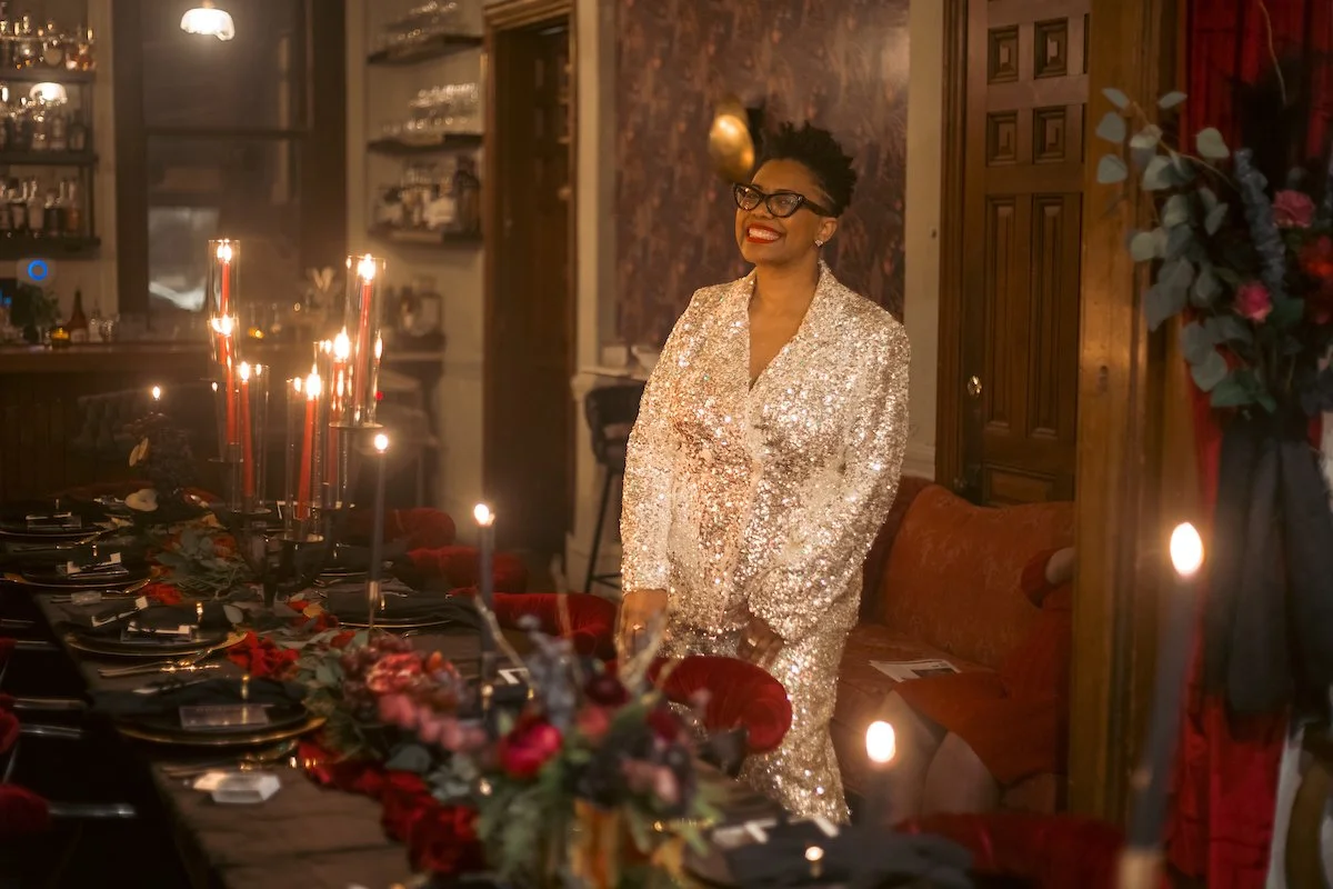 A woman in a sparkling gold dress standing next to a decorated dinner table with candles, smiling in a warmly lit room with wooden furniture and floral arrangements.
