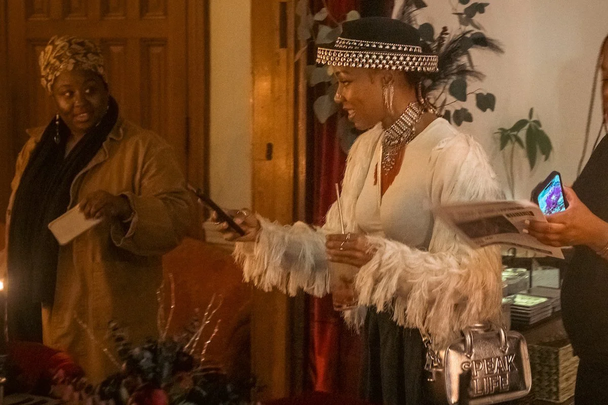Women at an indoor gathering; one woman in a white feathered coat and a hat with rhinestones holding a drink, another woman in a head-wrap and brown jacket, and a third woman in black holding a phone. There are plants and wooden furniture in the back