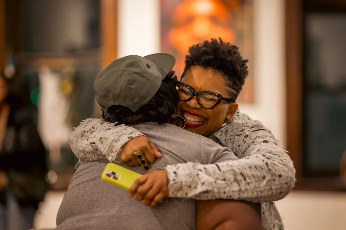 Two women hugging intimately, one smiling widely with eyes closed and wearing glasses, the other with her face hidden, holding a yellow phone.