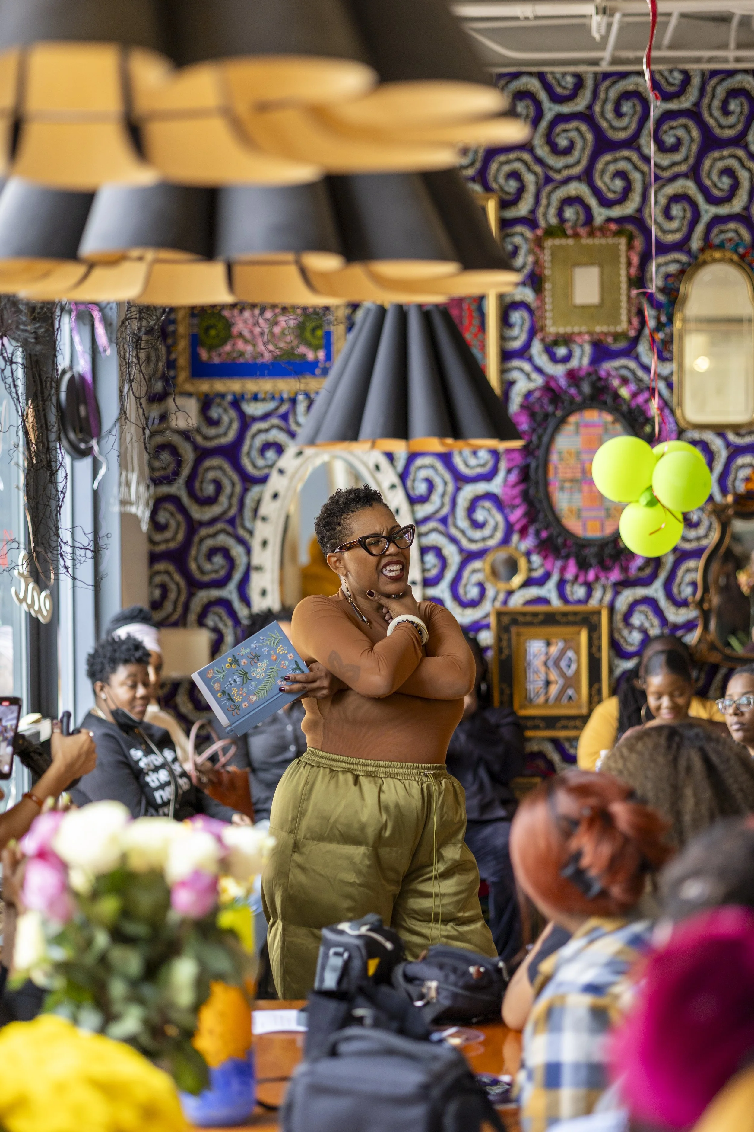 A woman speaking passionately at a gathering in colorful, decorated room with people watching attentively.