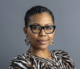 A woman with short curly hair, wearing glasses and earrings, dressed in a patterned top, posing against a gray background.