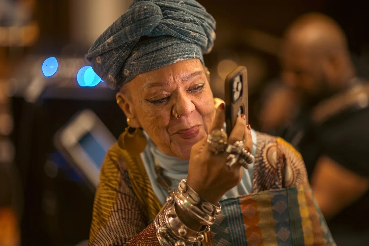 An elderly woman with a headwrap, earrings, and multiple silver bracelets, looking at her smartphone with a slight smile in a dimly lit setting.