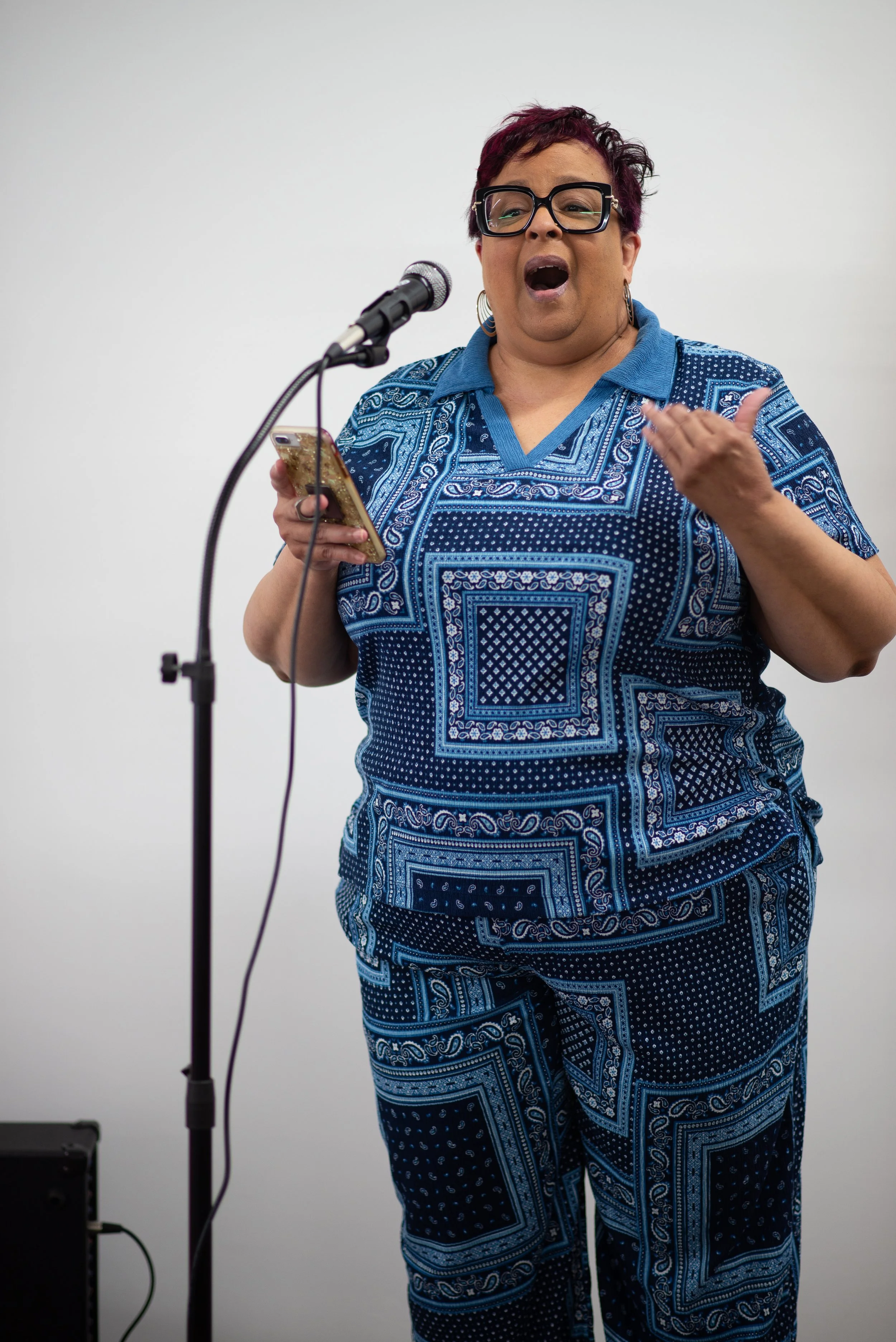 A woman with short dark hair and glasses, wearing a matching blue patterned outfit, stands in front of a microphone, speaking or singing while holding a smartphone.