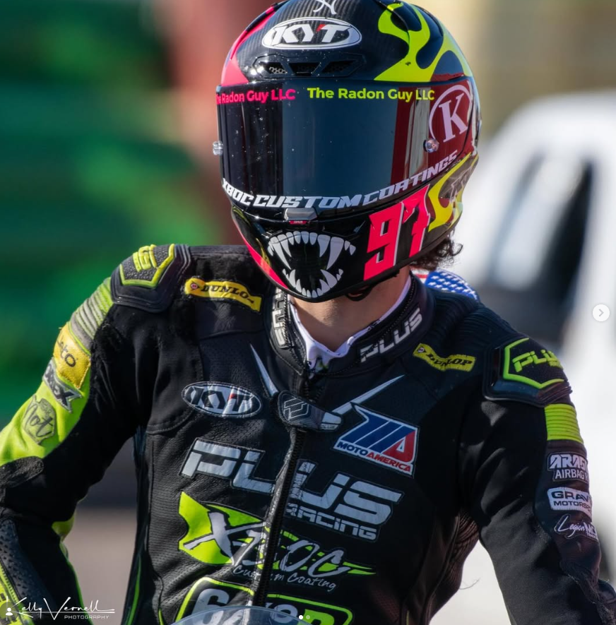 Motorsport racer wearing a colorful helmet and black racing suit with yellow accents, in a racing environment.