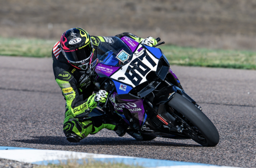 Motorcycle racing on a track, rider leaning into a turn, wearing a yellow, black, and pink helmet and a black and green racing suit.