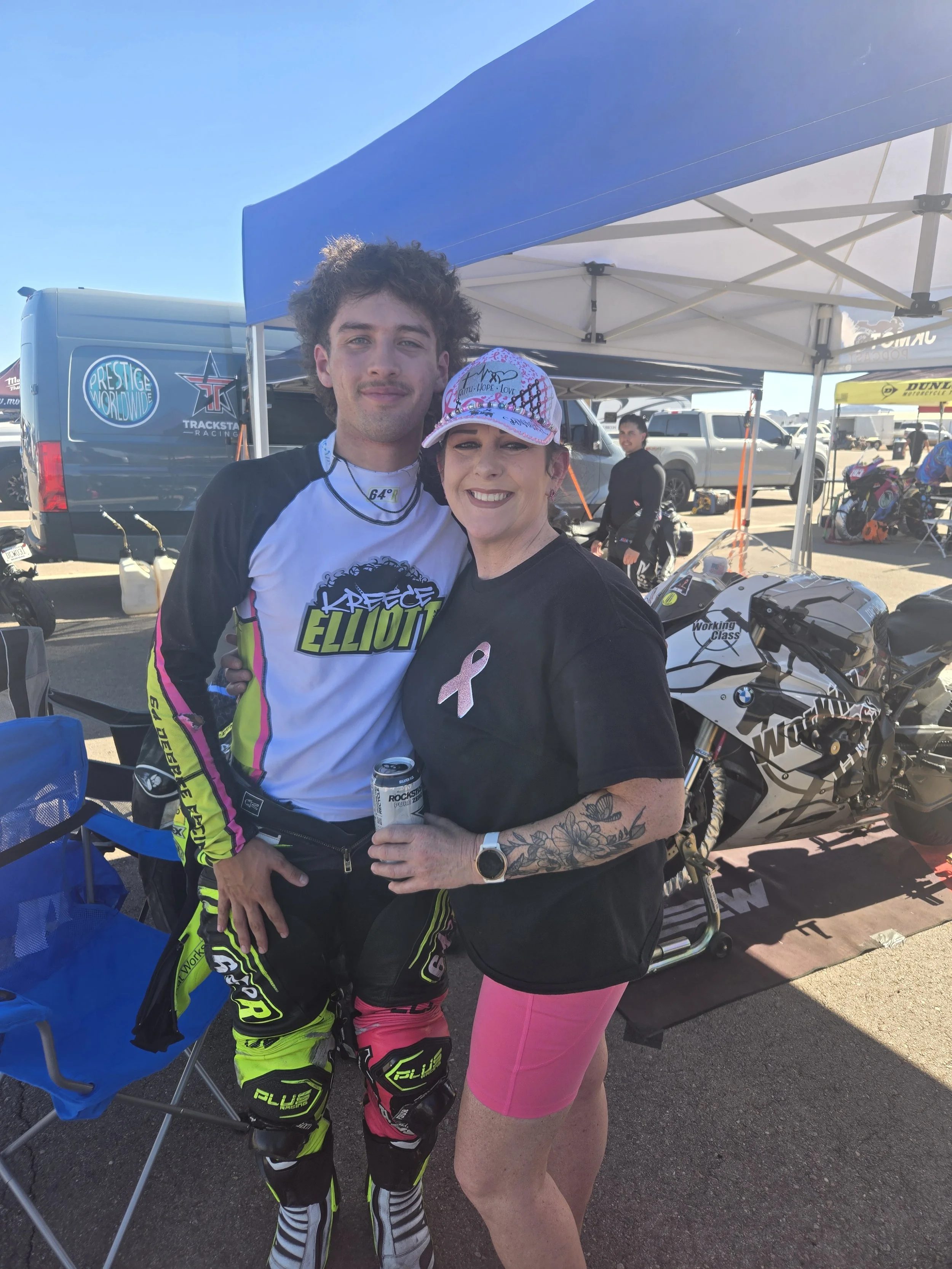 A man in racing gear and a woman in pink shorts and a black shirt with a pink breast cancer awareness ribbon pose together smiling outdoors at a racing event.