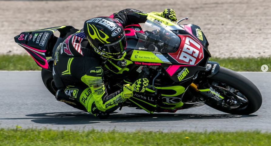 Motorcycle racer wearing a black and yellow racing suit and helmet, leaning into a turn at high speed on a race track.