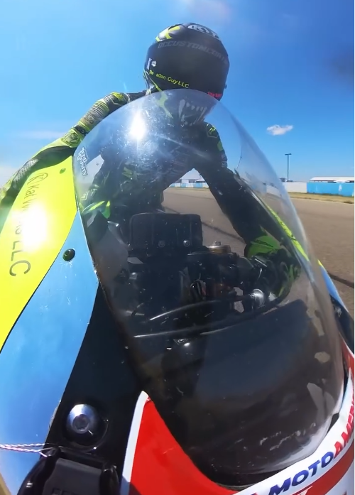 A motorcycle racer wearing a black helmet and a black and green racing suit, leaning forward on a motorcycle with a clear windscreen, on a racetrack with a yellow safety barrier and blue sky background.