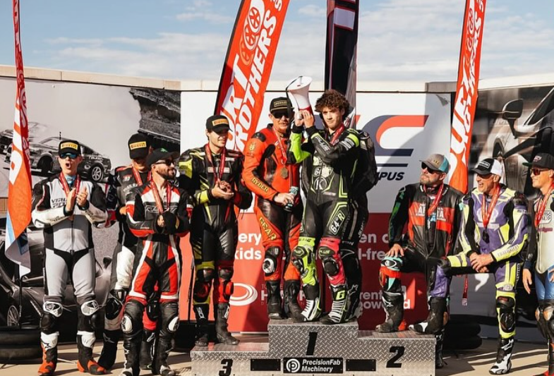 Motorsport winners on the podium celebrating with trophies, surrounded by other racers, in racing gear and helmets, with banners and promotional flags in the background.