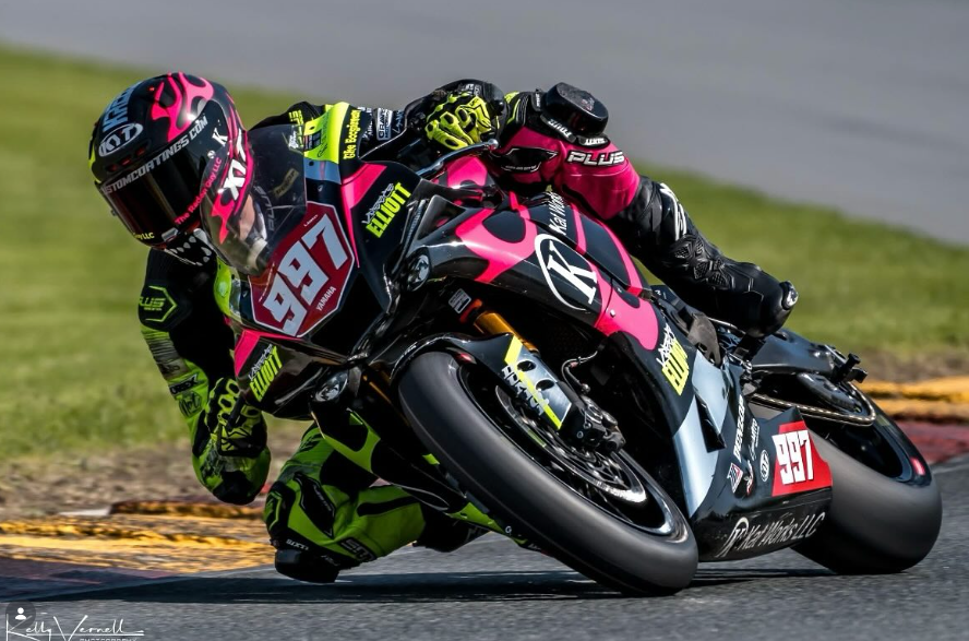 Motorcycle racer leaning into a turn on a racetrack, wearing a black and pink helmet and a black and pink racing suit, on a black motorcycle with racing decals and the number 987.