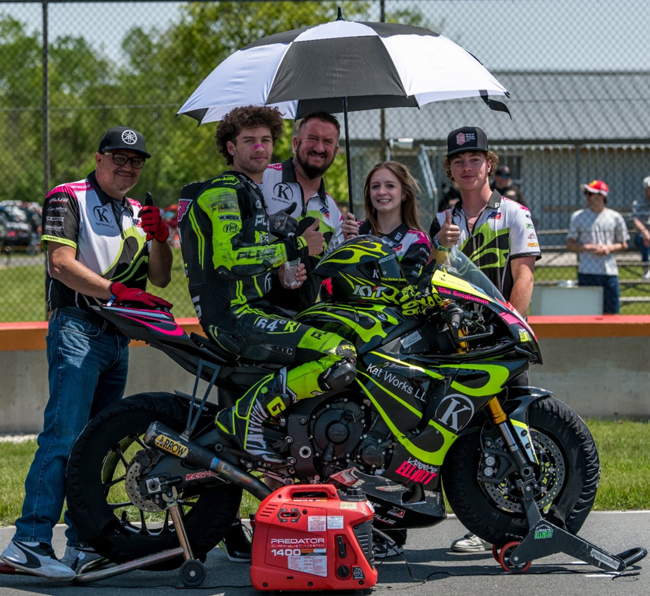 Group of five people at a racetrack, standing behind a racing motorcycle, holding an umbrella over the rider. The rider is seated on the motorcycle, wearing racing gear and a helmet. The group appears happy, giving thumbs up. There is a trash can and