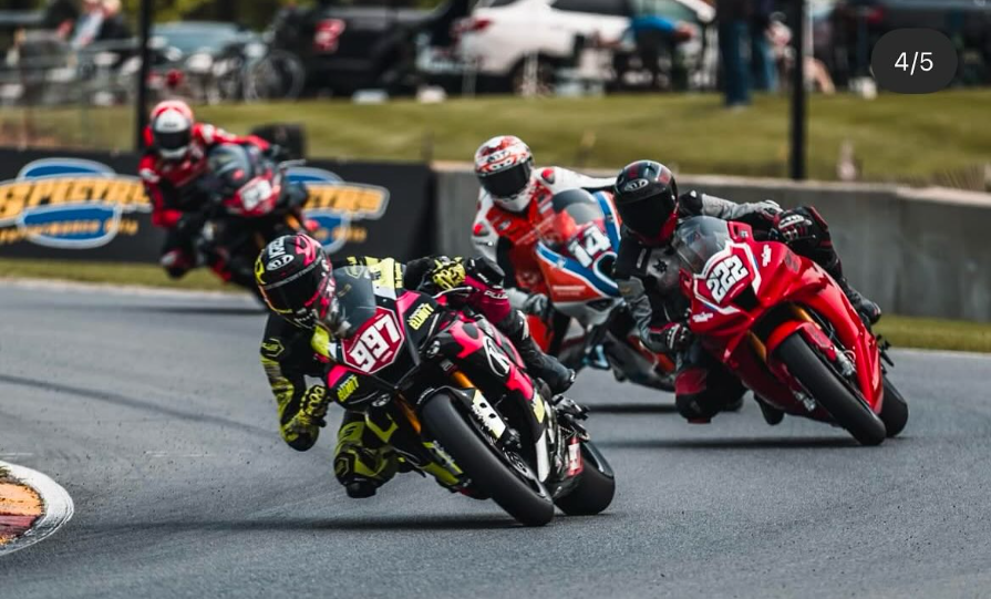 Four motorcycle racers leaning into a sharp turn on a race track, with motorcycles numbered 97, 222, 11, and 50. Spectators and cars are visible in the background.