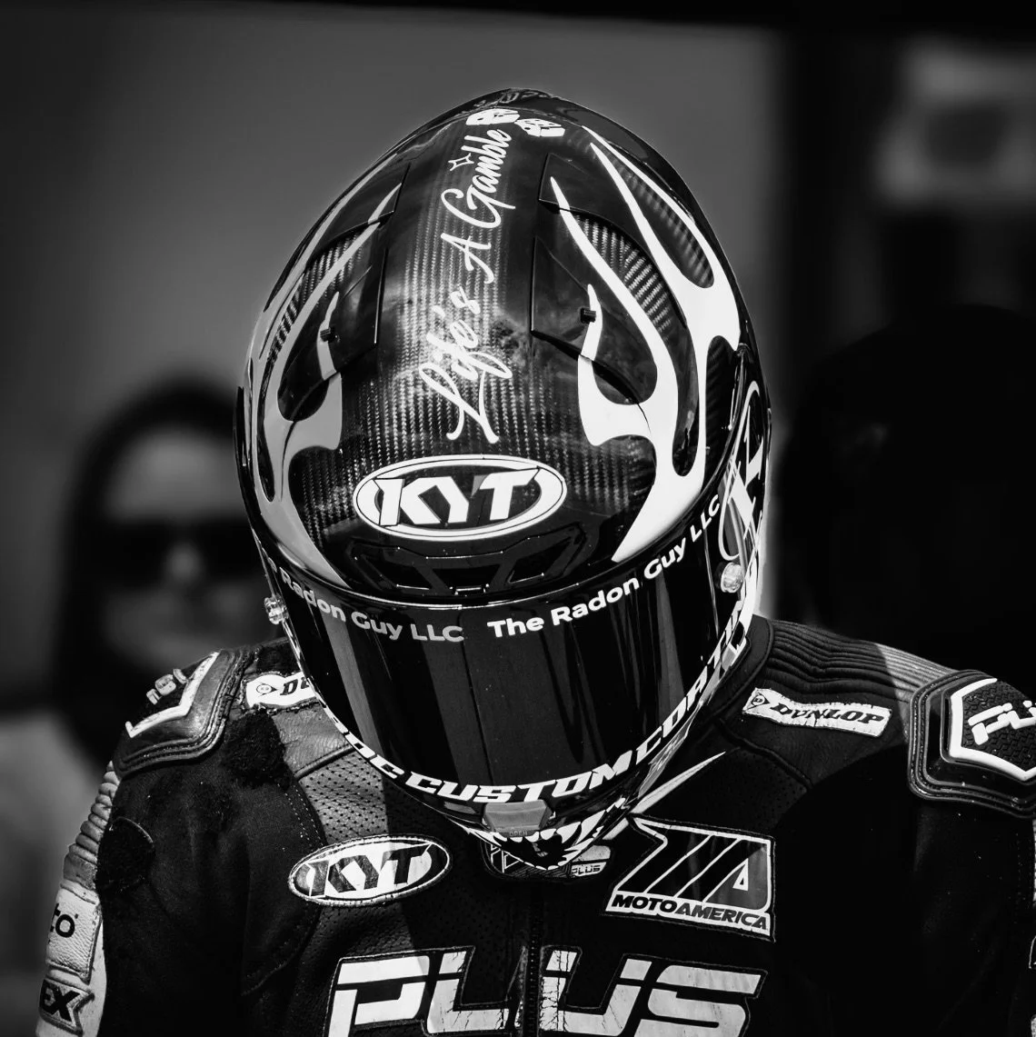 A professional motorcycle racer wearing a helmet and racing suit, packed with sponsor logos, in black and white.