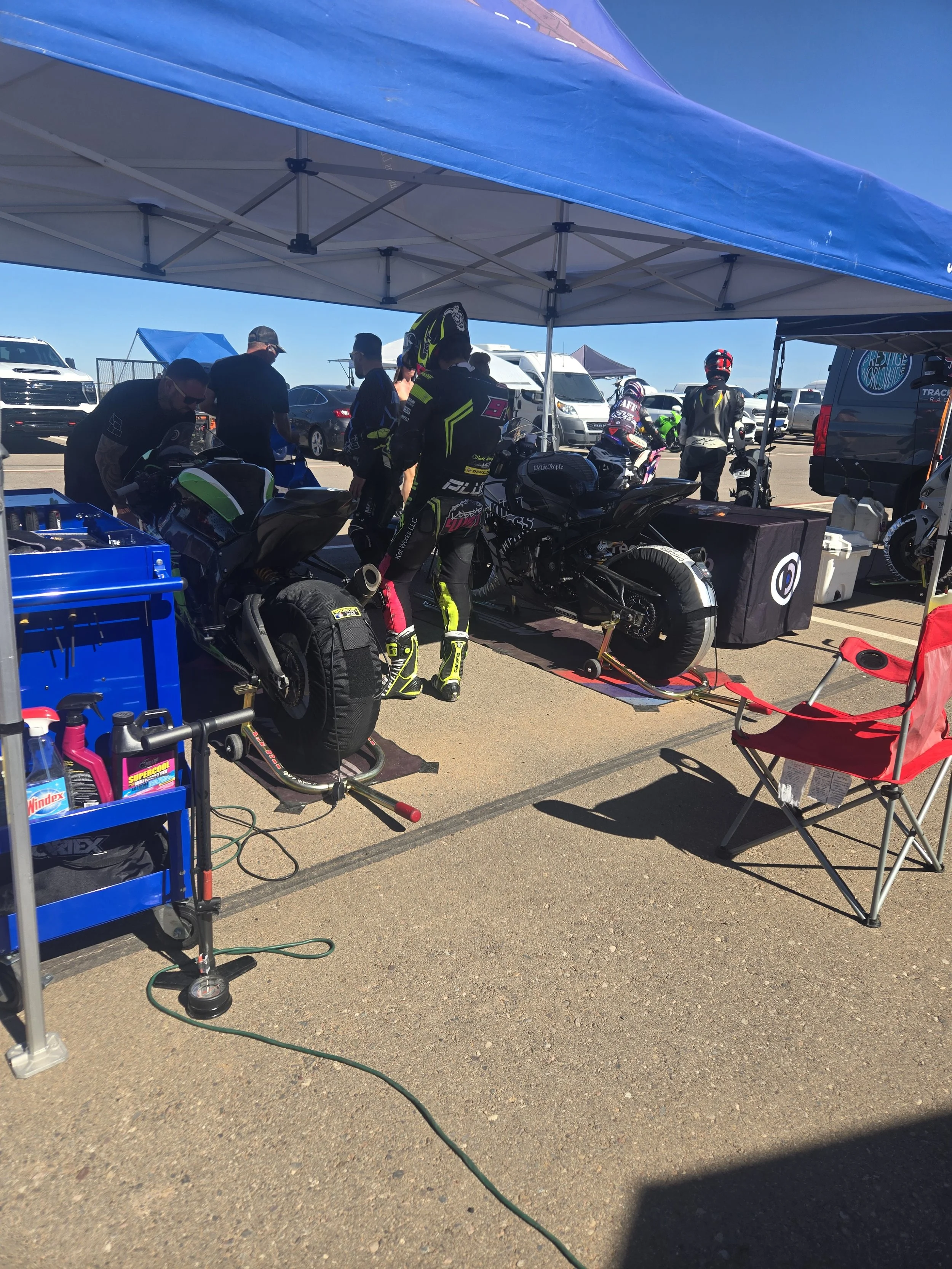 Motorcycle racing pit area with riders preparing bikes under a blue canopy, surrounded by tools, chairs, and vehicles.