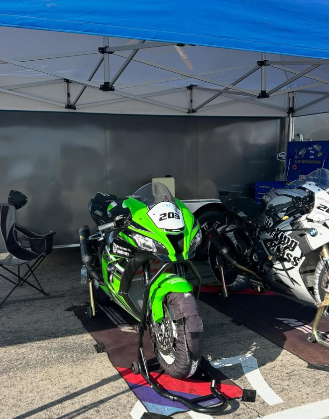 Two racing motorcycles under a blue canopy, one green and black, and the other white and black, are parked on race track mats with paddock stands. A folding chair is to the left.