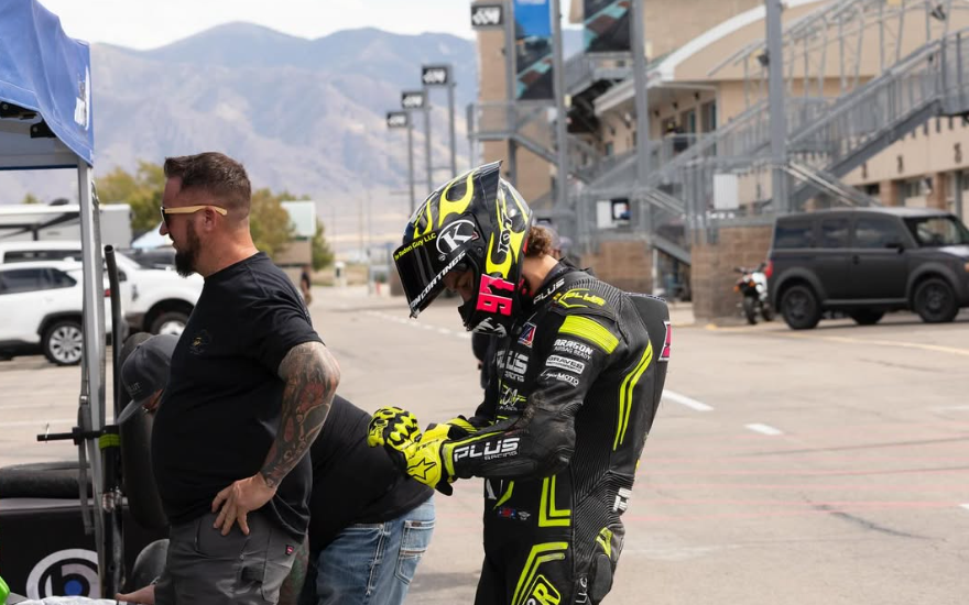 Motorsport rider in black and neon yellow gear with a helmet, signing an autograph in a parking lot with cars and mountain scenery in the background.