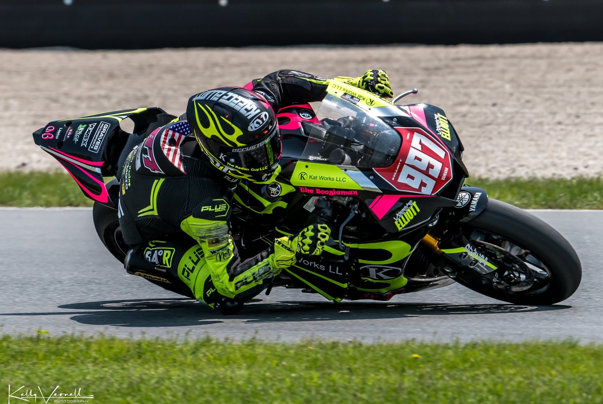 A motorcycle racer in black and neon yellow gear leaning into a turn on a race track, with the number 97 and various sponsor logos on the bike.