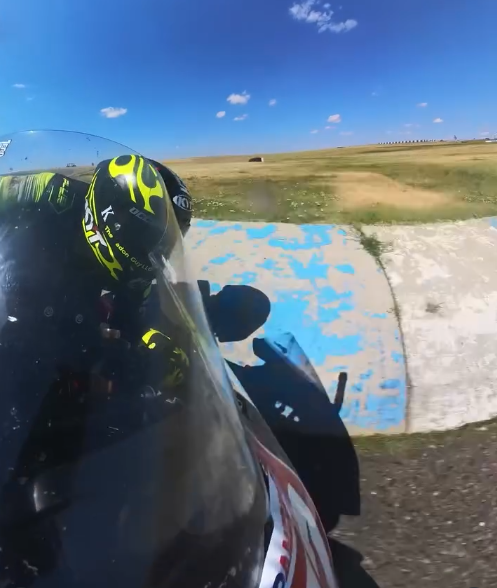 A person in a racing helmet and suit riding a motorcycle on a track with blue and white painted barriers, a grassy field, and a clear blue sky in the background.