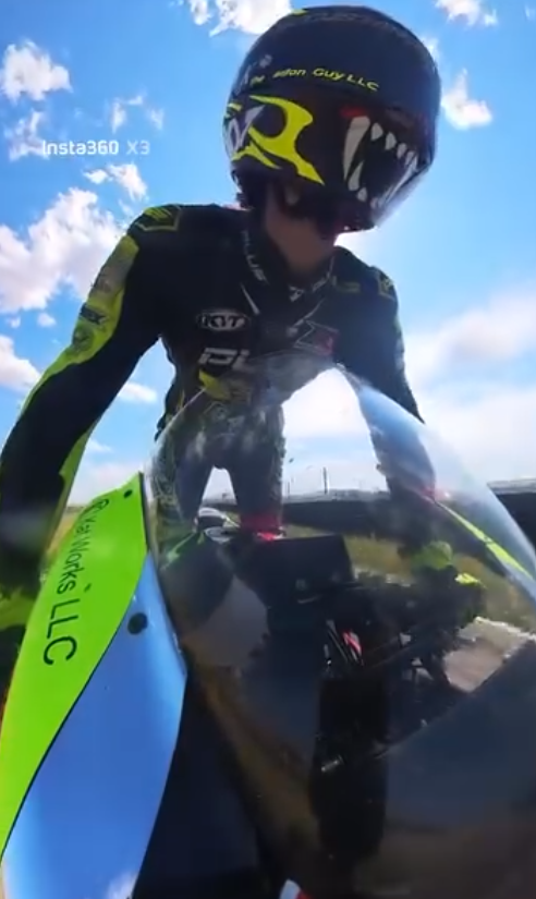 Motorcycle racer in a black and neon yellow suit and helmet, leaning over a black and green motorcycle, with a blue sky and clouds in the background.
