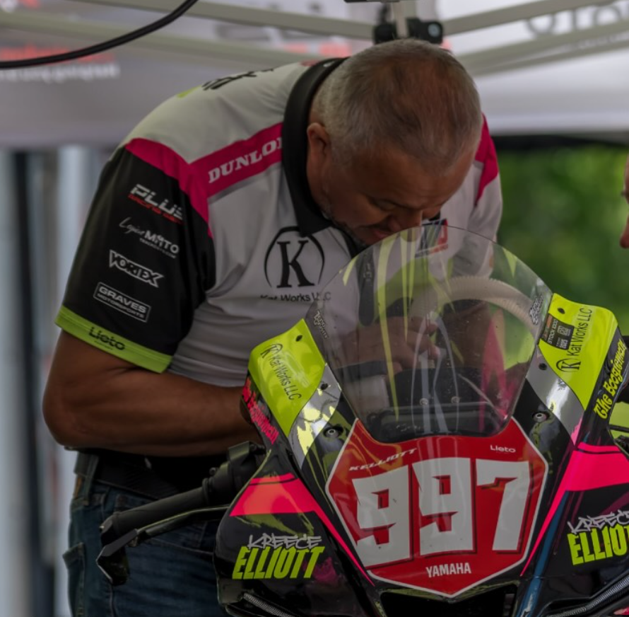 A man leaning over a racing motorcycle with the number 947, wearing a team shirt, in a pit area.
