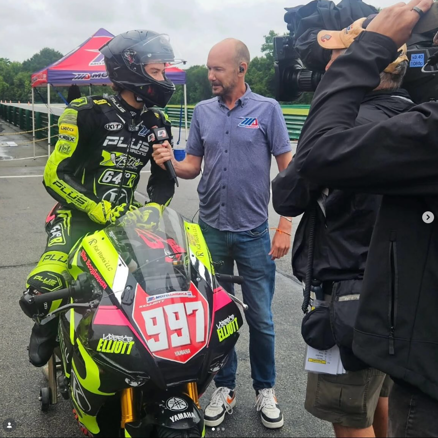 A motorcycle racer in a bright yellow and black racing suit being interviewed by a man with a microphone, beside a black and pink Yamaha race bike with the number 997. A cameraman recording the interview is partially visible.