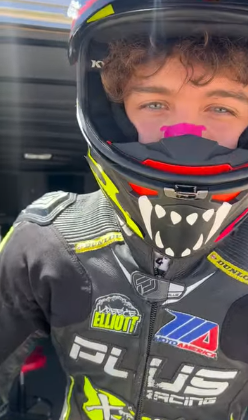 A young male race car driver wearing a full black racing helmet with red, yellow, and pink accents, and a racing suit with various sponsor logos, including Elliott and PluS Racing, standing in front of a vehicle with a blurred background.