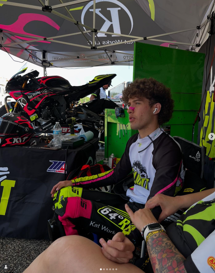 A young male motocross rider sitting in a pit area, wearing a racing suit with yellow and pink accents, earbuds, and with a helmet on the table nearby. The area is covered by a tent with racing branding, and there is a motorcycle and gear in the back