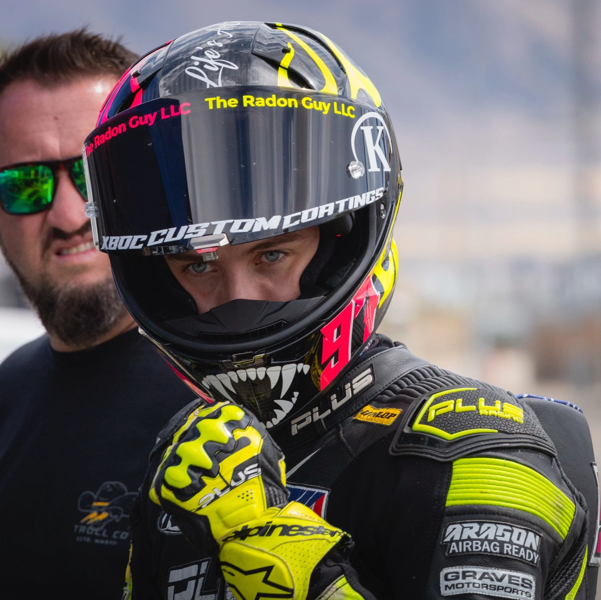 A race car driver wearing a black and yellow racing suit and helmet, adjusting their gloves, with another person in sunglasses in the background.