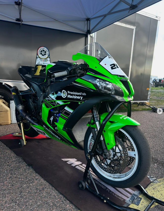 Green and black racing motorcycle under a tent at a race track, with front wheel secured in a paddock stand, and a sponsorship decal visible.