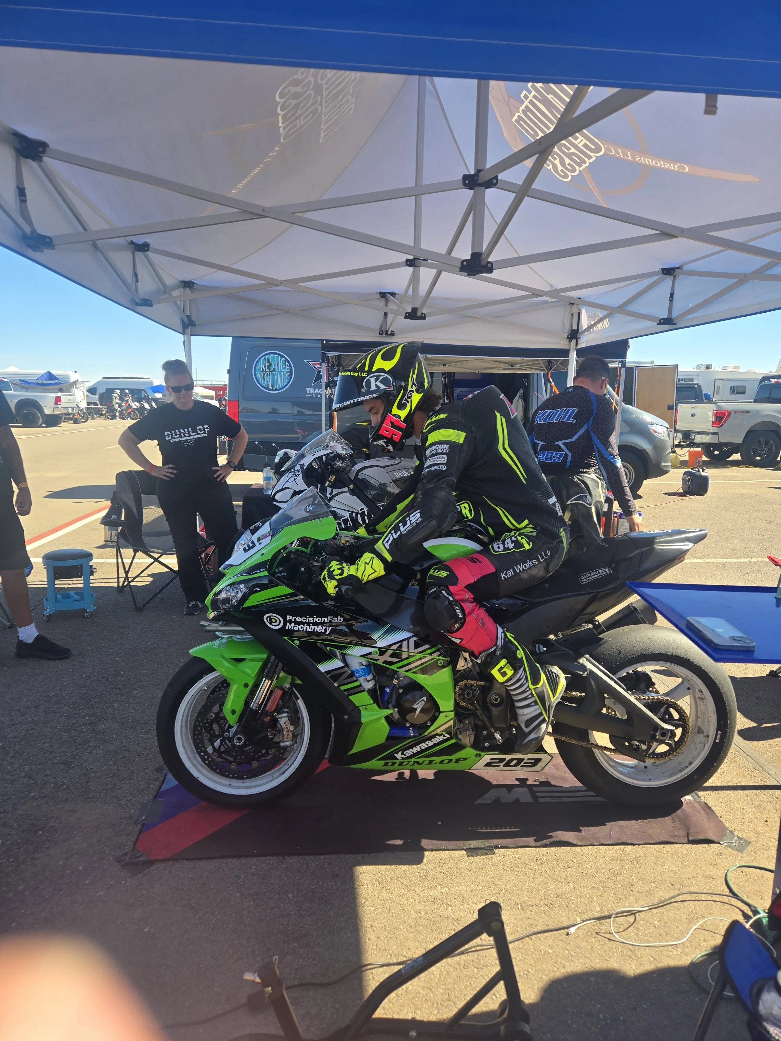 A motorcycle racer in racing gear sits on a green Kawasaki racing motorcycle under a white tent at a racetrack, with team members and support vehicles in the background.