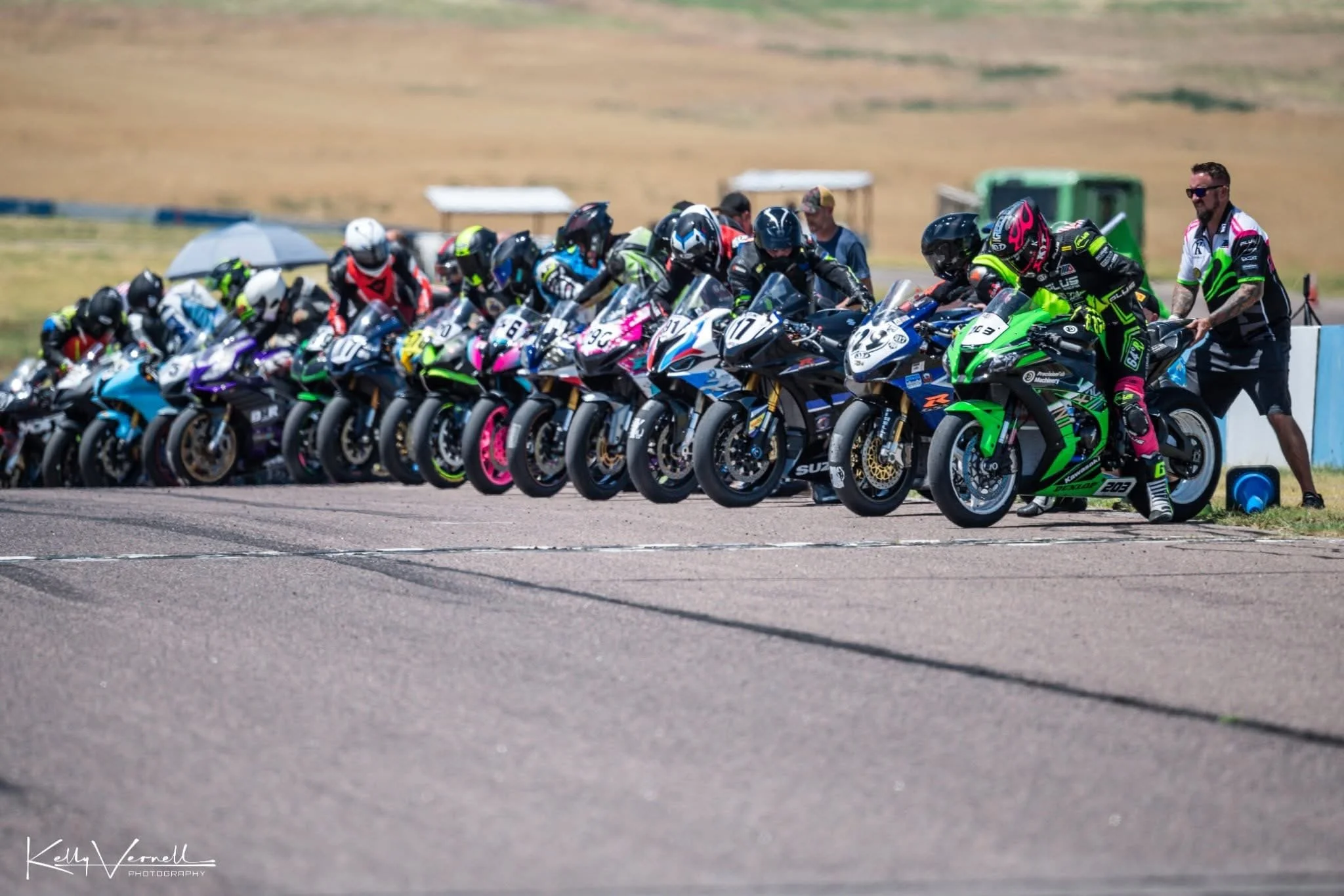 Line of motorcycle racers at the starting grid during a race event, with race officials preparing the bikes.