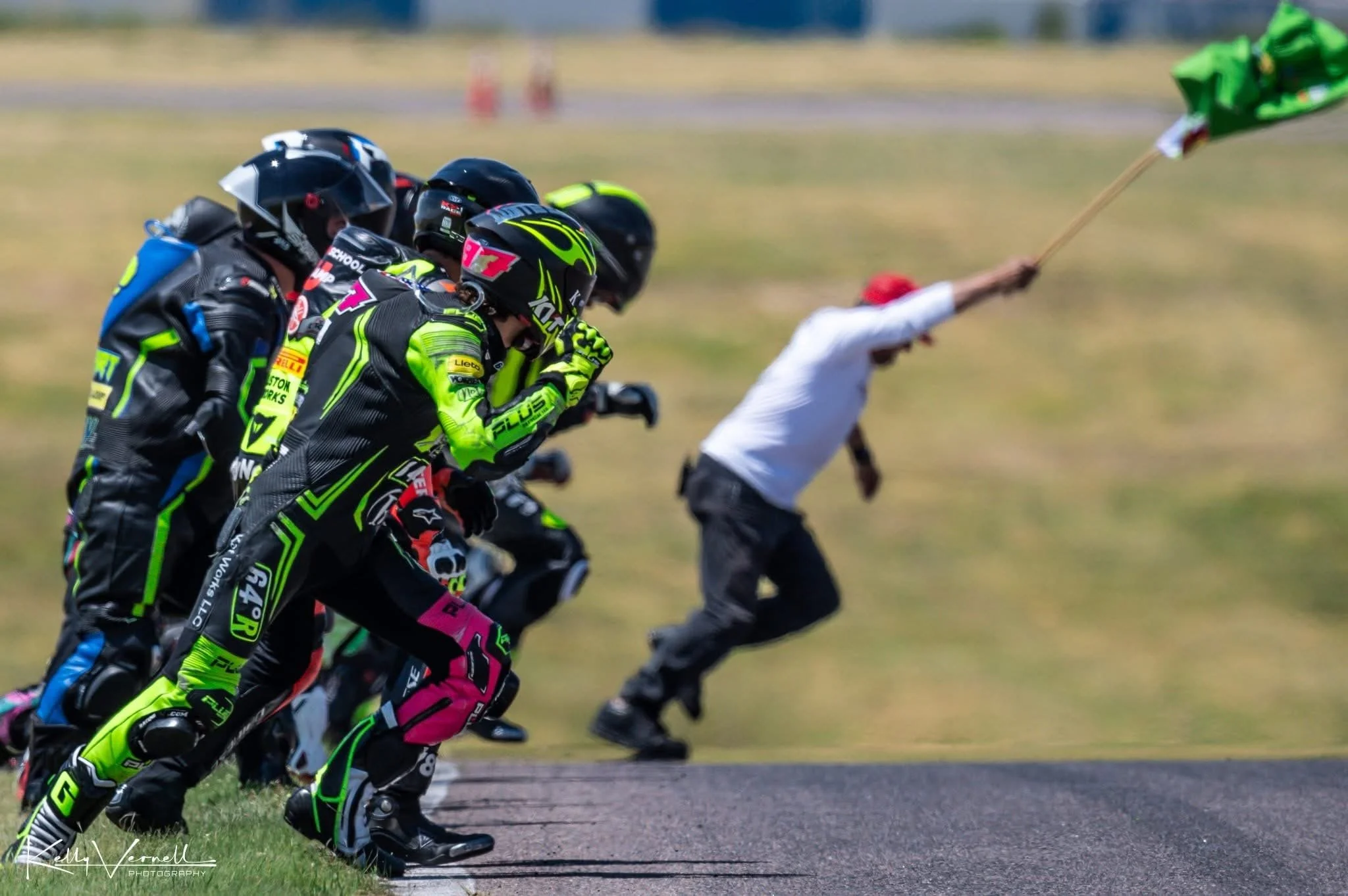 Motorcycle racers preparing at the starting line on a race track, with a track official waving a green flag to signal the start.