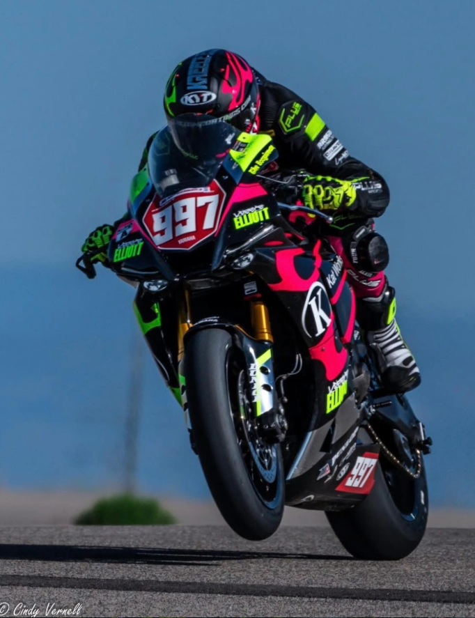 Motorcycle racer in black, neon yellow, and pink racing gear. The motorcycle features black with bright pink accents, multiple sponsor stickers, and the number 997 on the front. The rider is leaning into a turn on a race track with a blue sky backgro