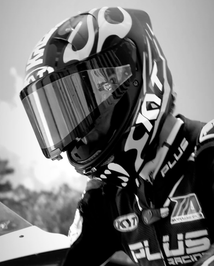 A person in full racing gear, including a helmet with a visor, is preparing for a motorcycle race outdoors. The image is in black and white.