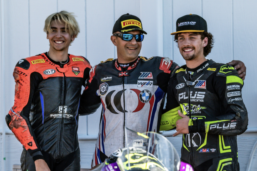 Three race car drivers in racing suits and caps smiling and standing together against a white wall.