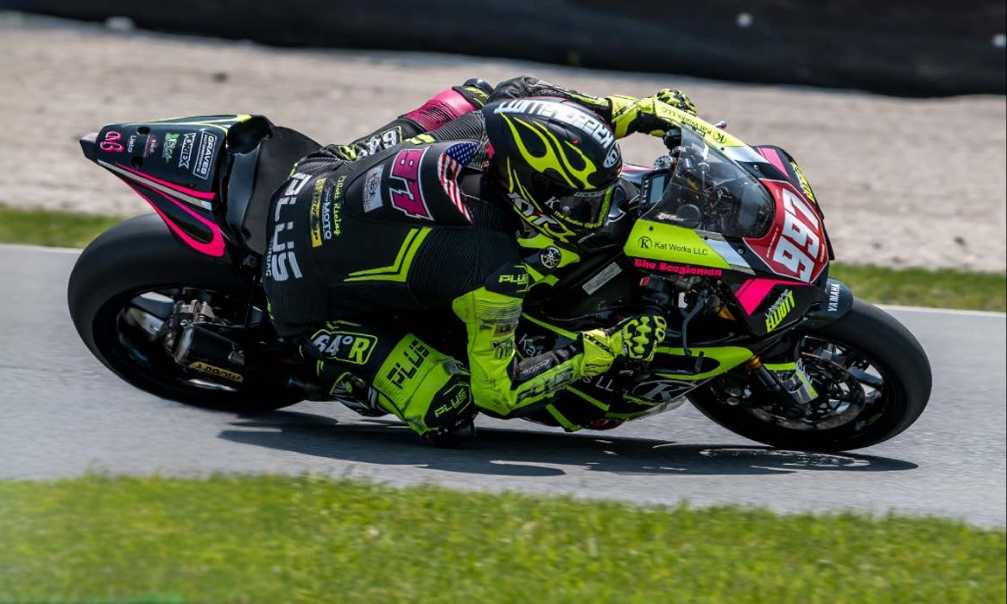 Motorcycle racer leaning into a turn on a racing track, dressed in a colorful helmet and suit with neon yellow and black accents, on a black and pink motorcycle with various sponsor logos.