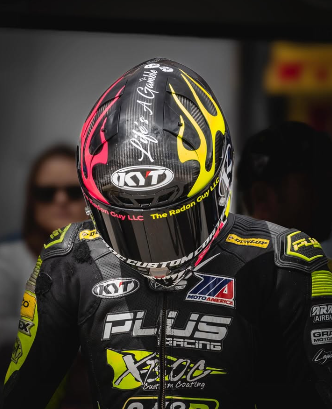 A motorcycle racer wearing a black racing suit with yellow accents and a matching helmet with pink and yellow designs, signature graphics, and sponsor logos, standing in a garage or paddock area.