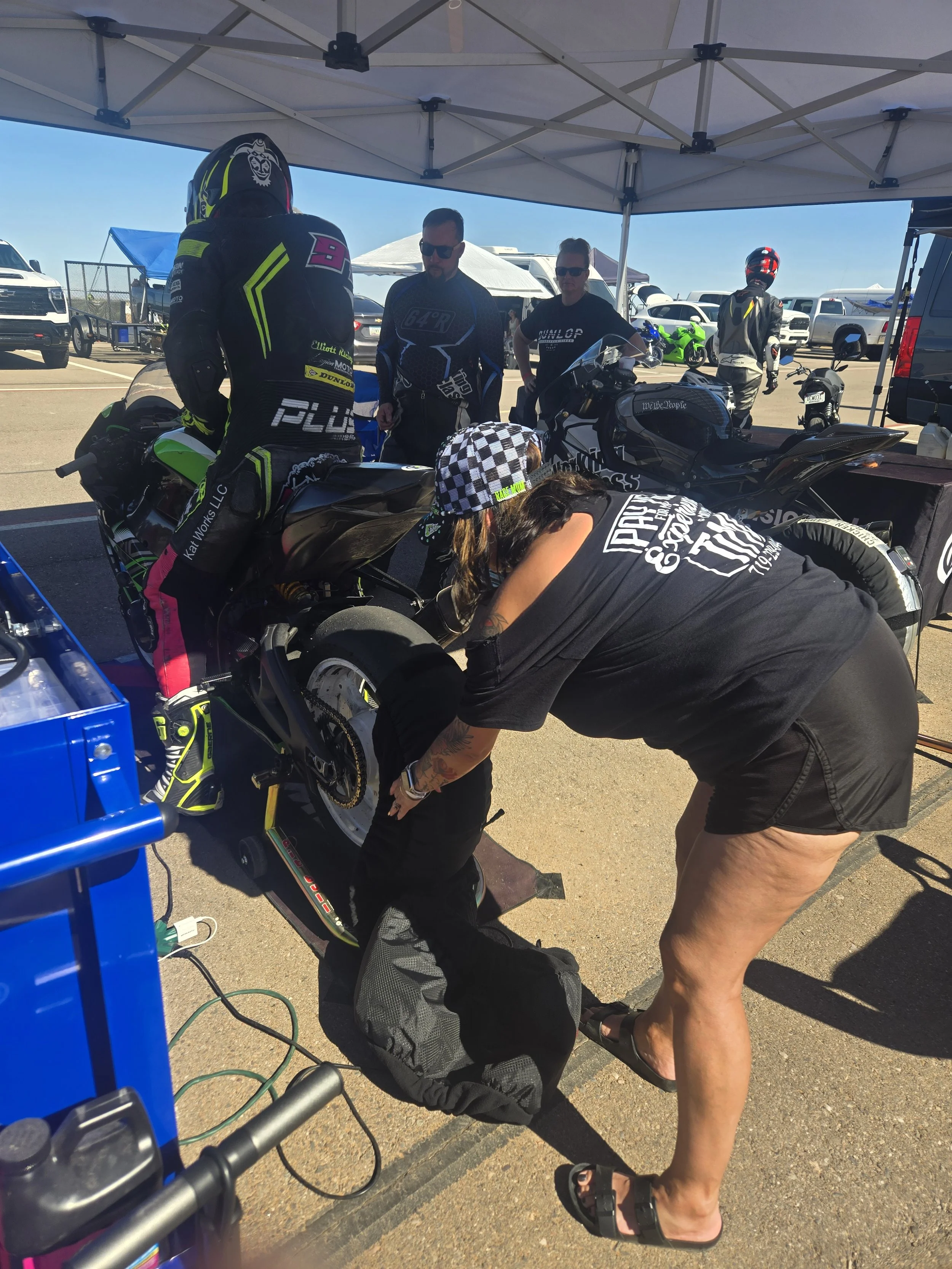 A woman in a checkered cap working on a black motorcycle under a tent at a racetrack, with several people observing in the background.