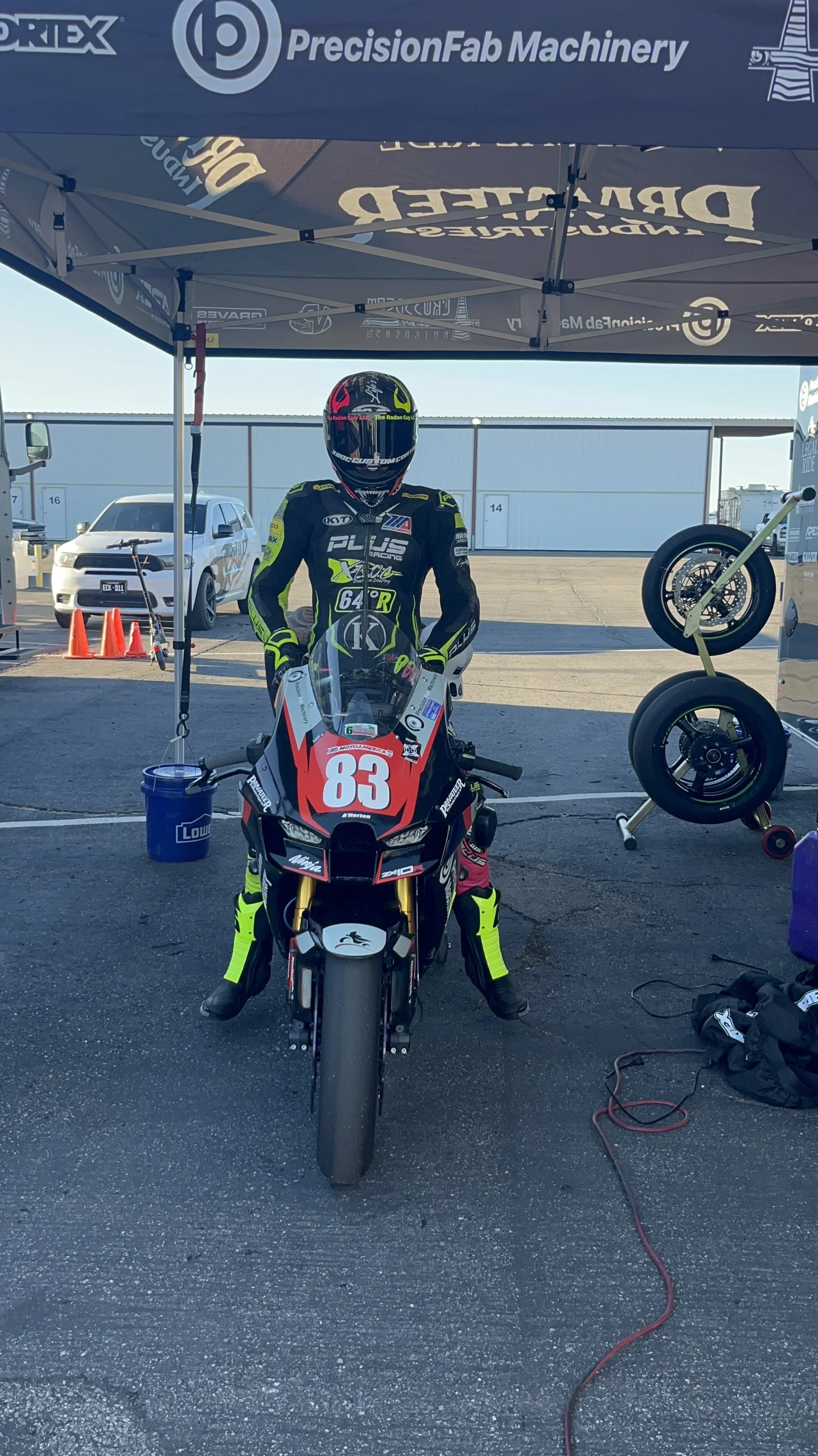 Motorcycle racer in black and neon yellow riding suit standing behind a red and black race bike with the number 83 on it, under a canopy at a racing paddock.