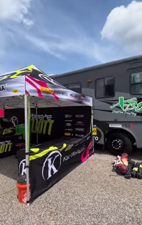 A racing team setup beside a large black RV with green branding, including a tent with sponsor decals, a table, and equipment on the ground, under a partly cloudy blue sky.