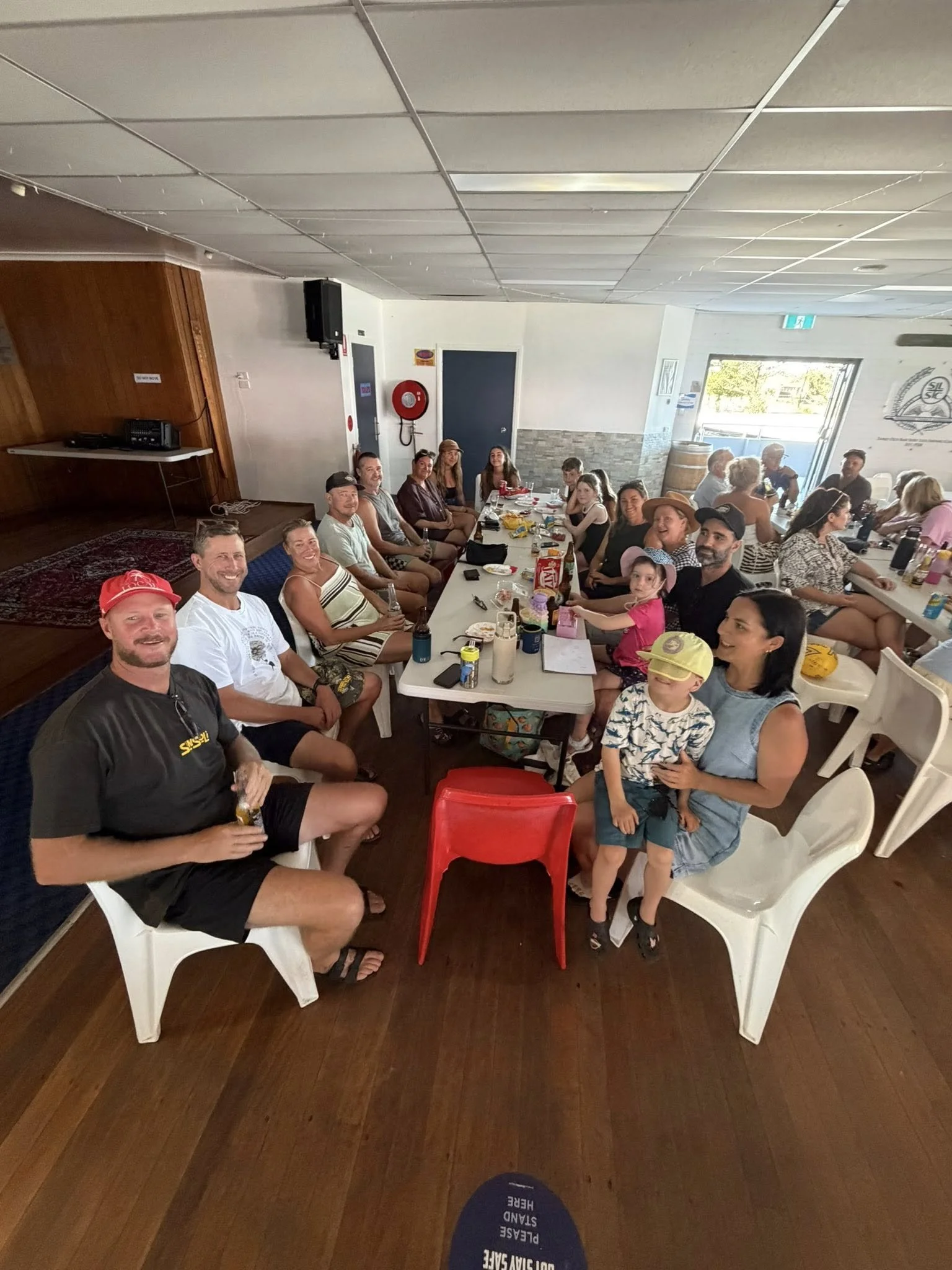A large group of people sitting around a long table inside the Surf Club. The table has snacks, drinks, and personal items. People are smiling and looking at the camera.