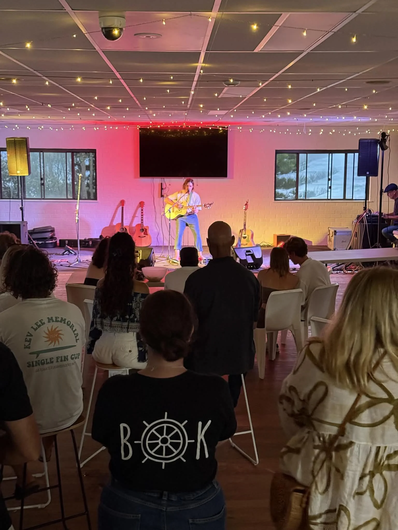 Indoor event space with a performer playing guitar on stage, surrounded by guitars, audience seated and standing, string lights hanging from the ceiling, large screen at the back, two large windows on sides, and a person on the right operating audio 