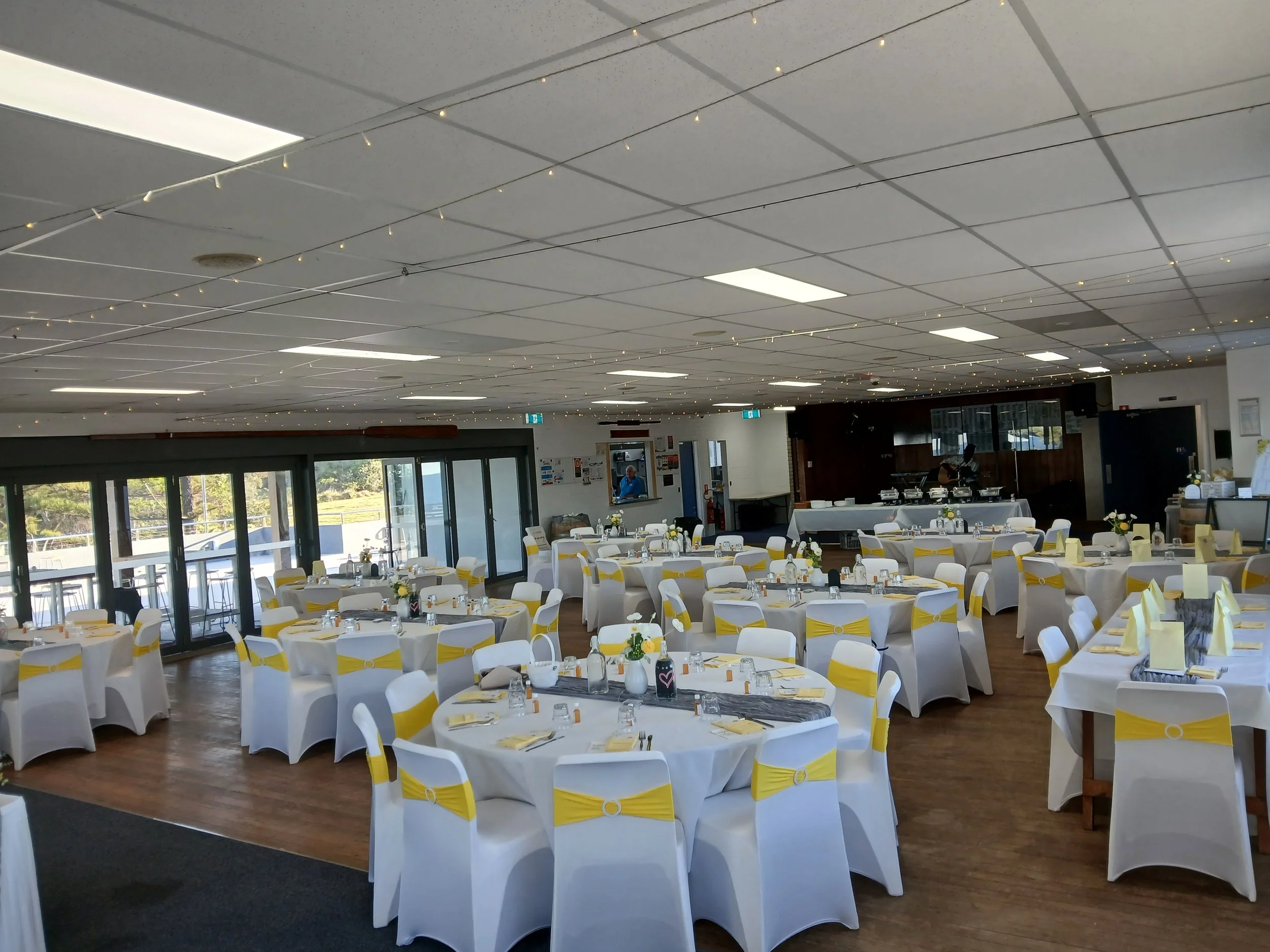 The function room set-up for a wedding showing individual round tables of 10 seats.  To the left are the concertina doors to the deck. Towards the back is a table set for a buffet dinner. 